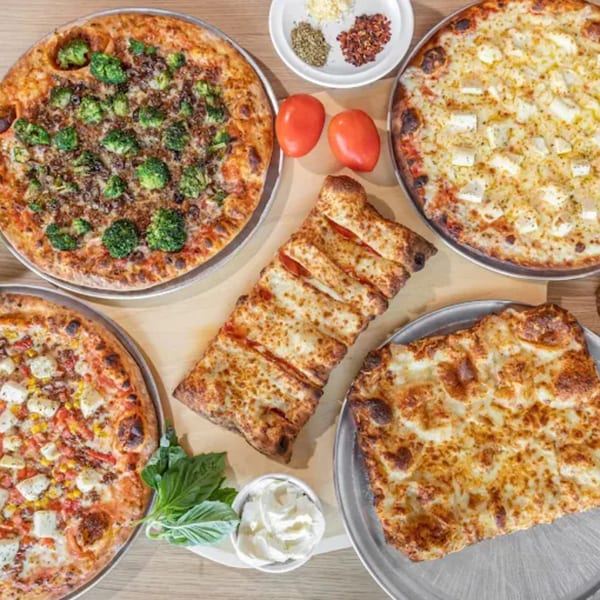 Four Via Capri Pizzeria pizzas, a calzone, tomatoes, basil, and spices on a wooden table.