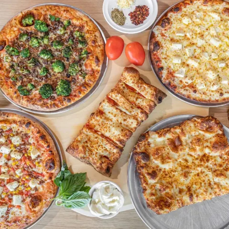 Four Via Capri Pizzeria pizzas, a calzone, tomatoes, basil, and spices on a wooden table.